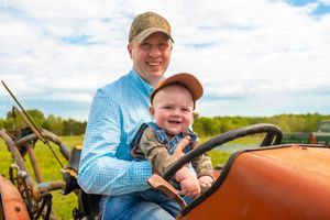 A person wearing a blue shirt and baseball cap operates an orange tractor in a sunny rural field.