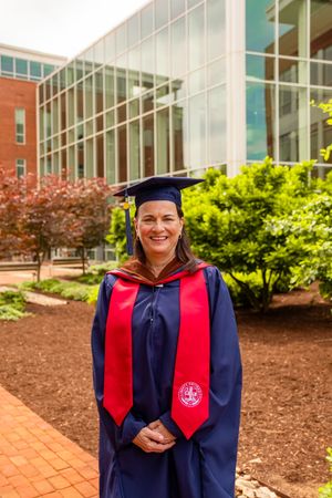 Graduate in navy blue cap and gown with red stole stands smiling outdoors near brick walkway and landscaping.