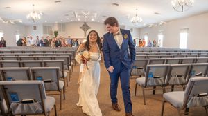 A couple walks down the aisle in a bright indoor venue with gray chairs and crystal chandeliers overhead.