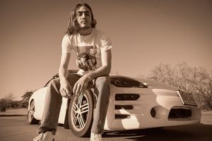 A sepia-toned image showing someone sitting on the hood of a modern sports car in an outdoor setting.