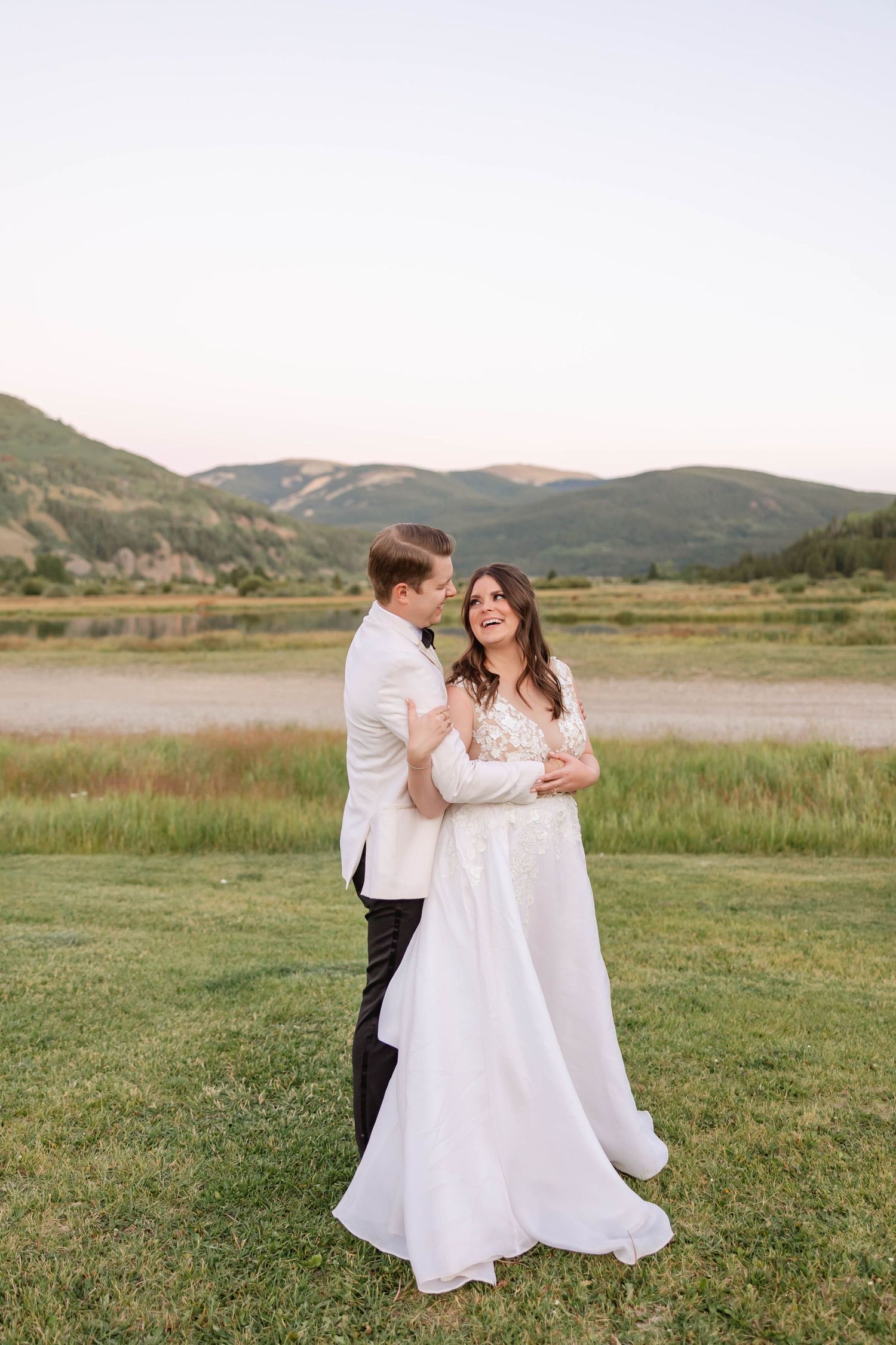 Wedding couple embraces on grassy field with mountain landscape in background.