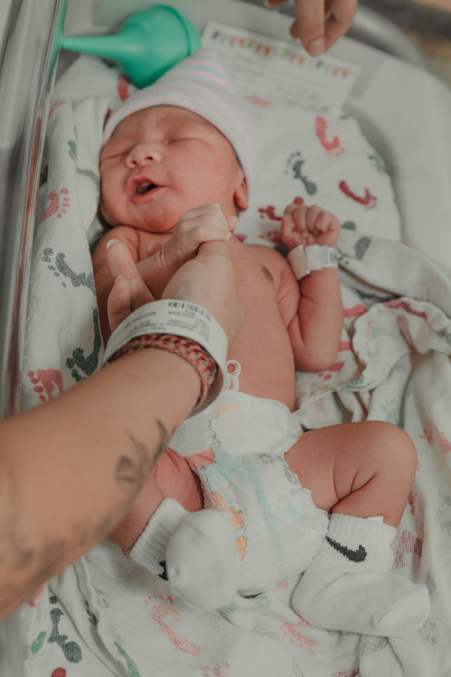 Hand tending to newborn baby wearing patterned blanket in hospital bed.