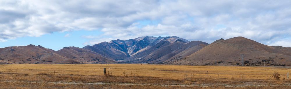 Wide panoramic view of brown plains stretching toward snow-dusted mountain ranges under a partly cloudy sky.