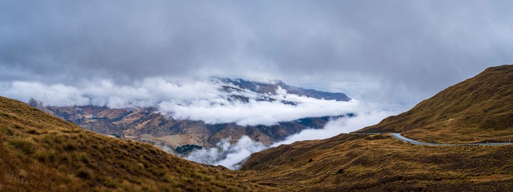 Panoramic view of snow-capped mountains shrouded in dramatic clouds above a rugged highland valley landscape.