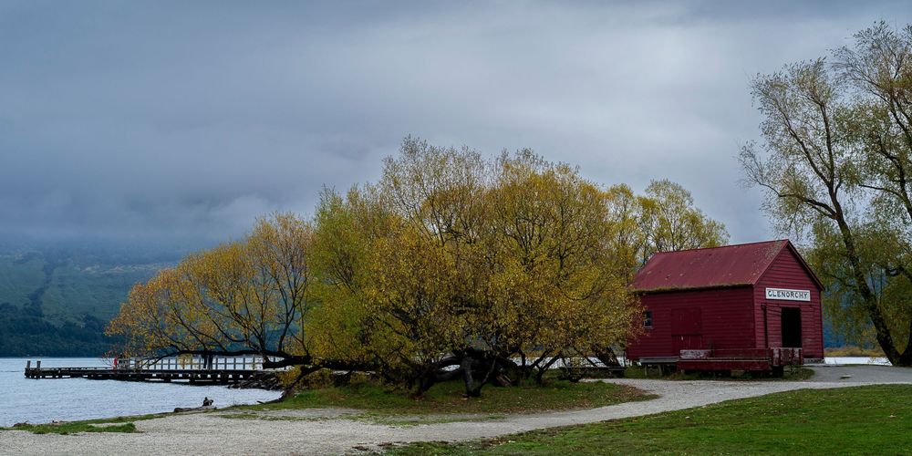 Red boathouse beside golden willow trees on a calm lakefront under overcast skies in New Zealand.