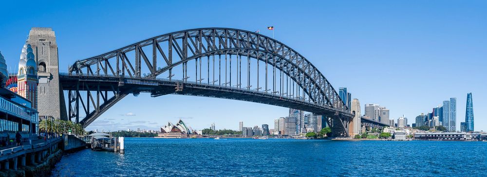 Panoramic view of Sydney Harbour Bridge with Opera House and city skyline reflecting on blue waters on a clear day.