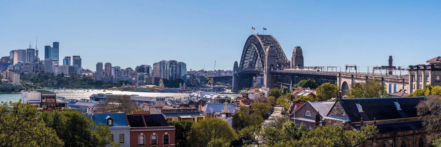 Panoramic view of Sydney Harbour Bridge and city skyline on a clear sunny day, with historic rooftops in the foreground.