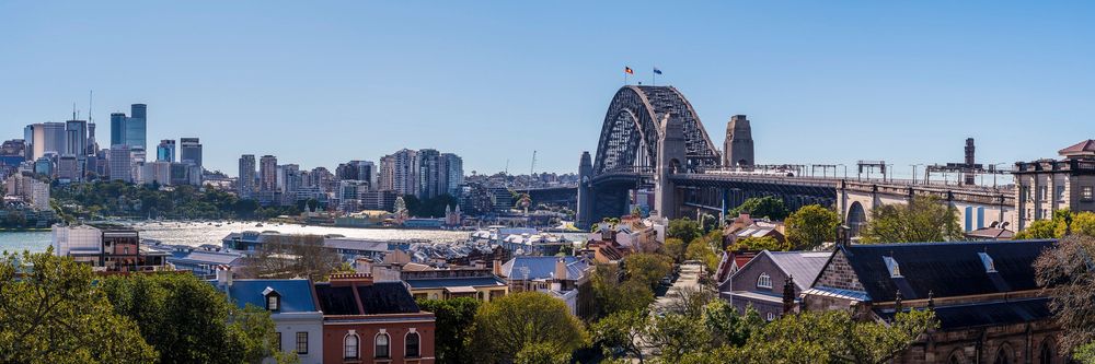Panoramic view of Sydney Harbour Bridge and city skyline on a clear sunny day, with historic rooftops in the foreground.
