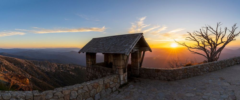 A panoramic sunset view from a stone lookout shelter atop a mountain with a dead tree silhouette.