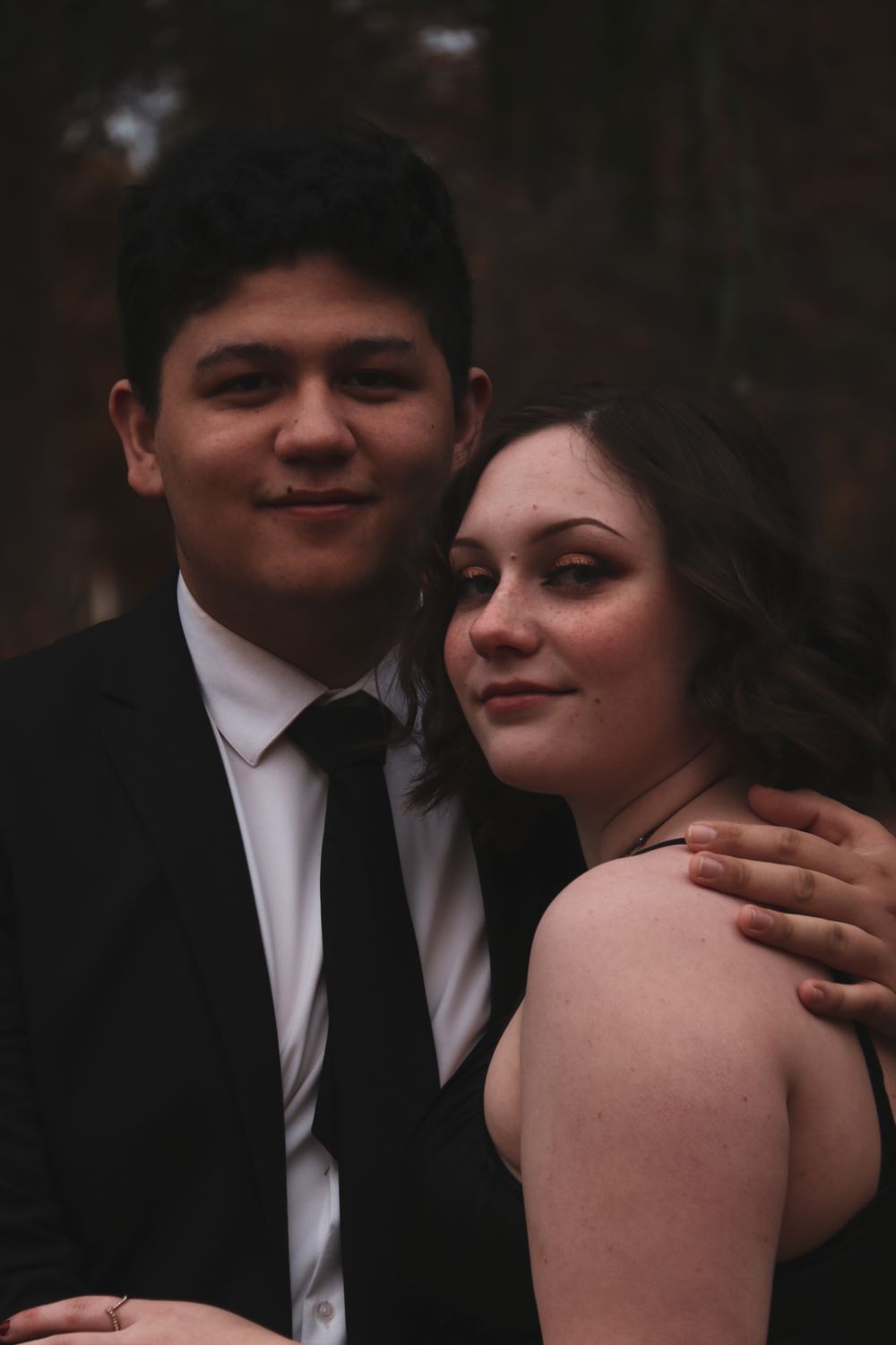 Couple posing together in formal attire, man in black suit with tie and woman in black dress, smiling at camera.