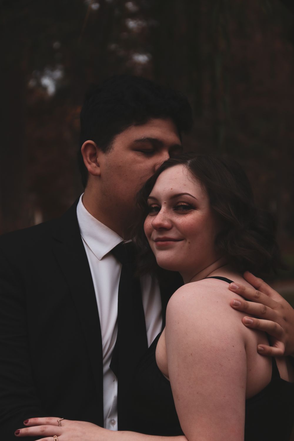 Man in black suit kissing woman's temple while embracing her from behind in a forest setting.
