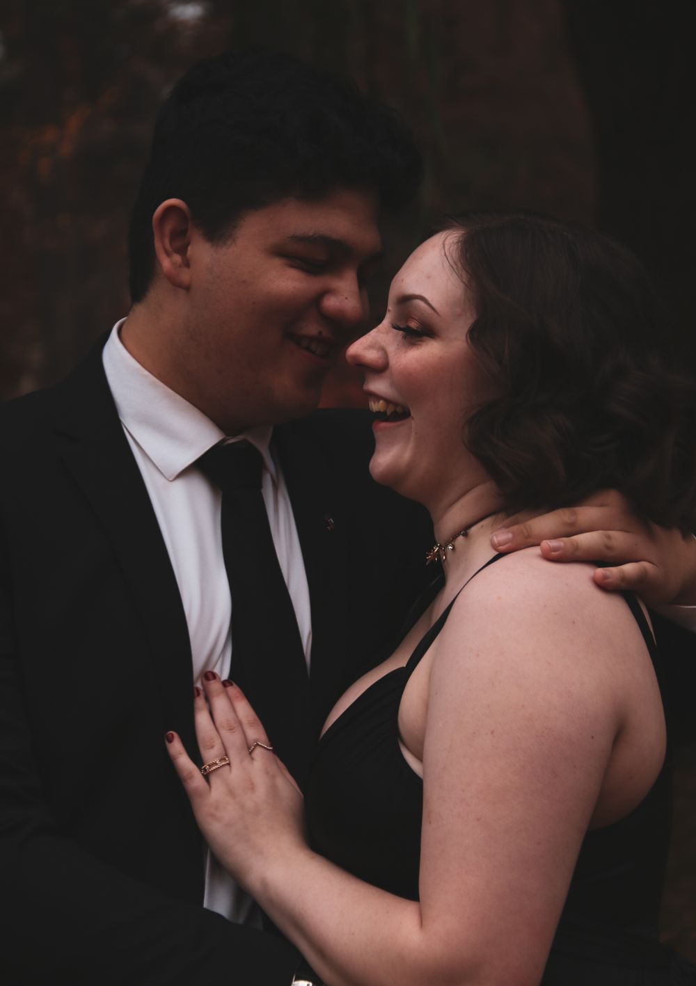 Couple laughing and embracing outdoors, man in black suit and woman in black dress sharing a joyful moment.