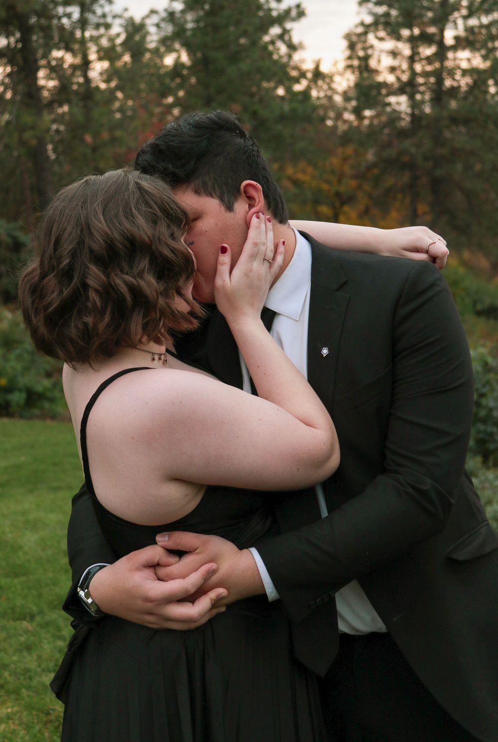 Couple kissing passionately outdoors at dusk, woman in black dress embracing man in suit.