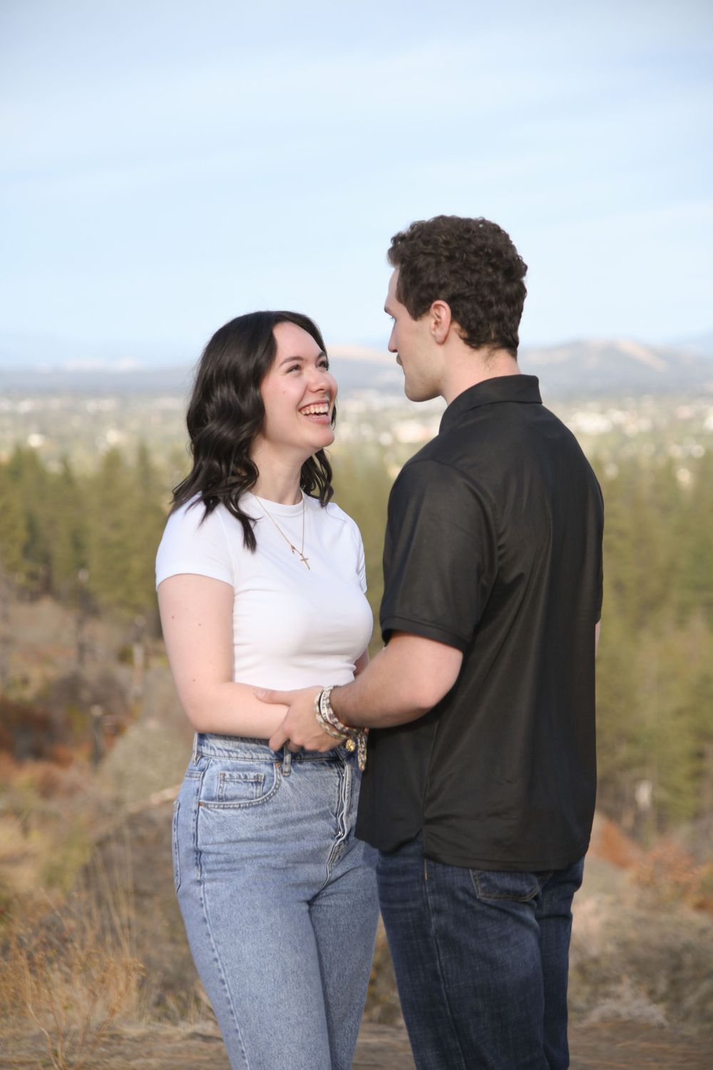 Smiling couple holding hands on a scenic overlook with a city skyline and blue sky behind them.