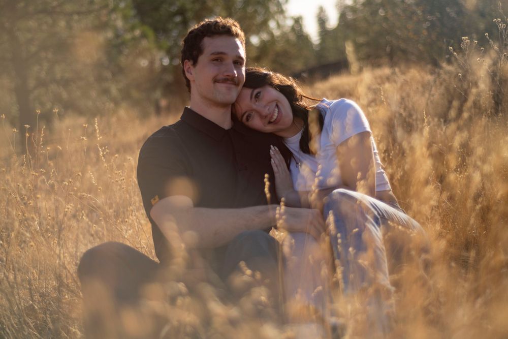 Happy couple sitting together in a golden sunlit field, woman resting head on mans shoulder, smiling warmly.