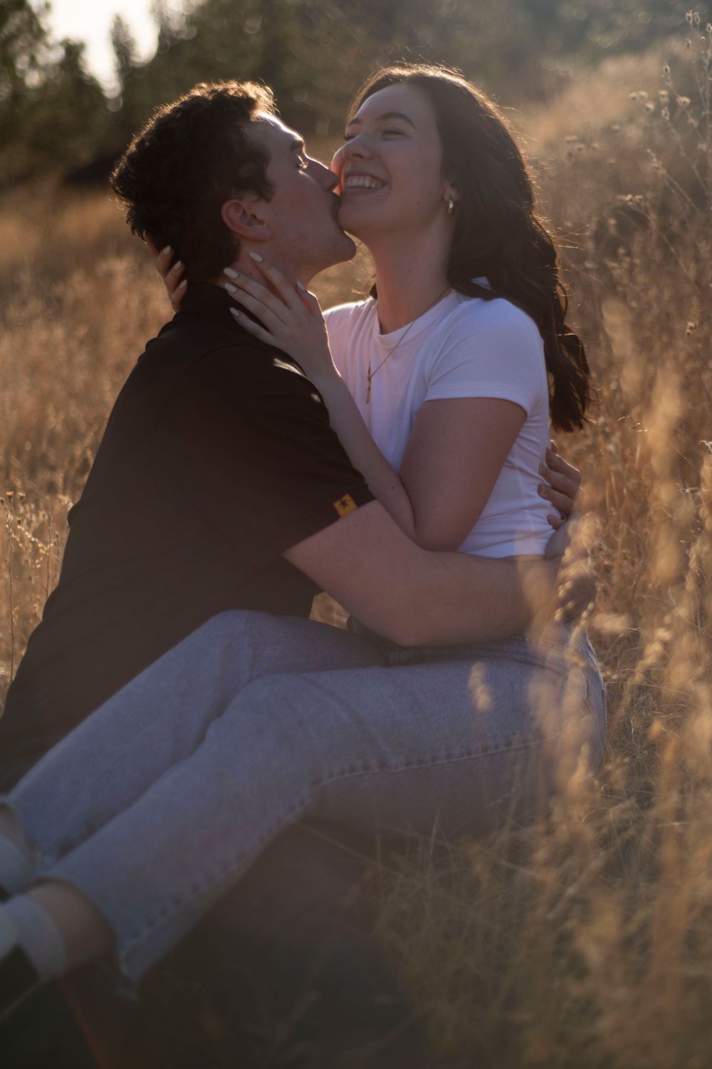 Romantic couple embracing in sun-drenched meadow, woman laughing as partner leans in to kiss her.