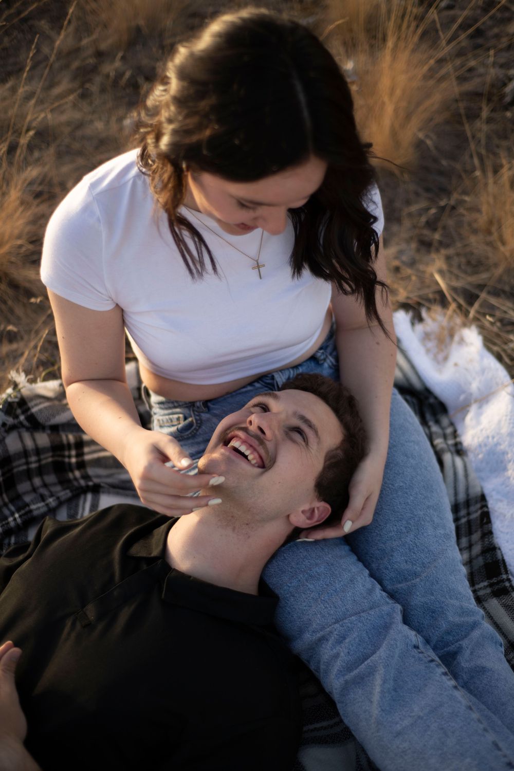 A laughing couple enjoys a playful moment on a blanket in a sunlit golden field outdoors.