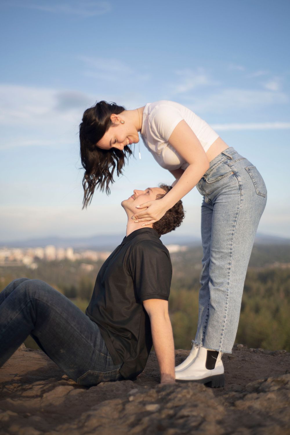A woman in a white crop top leans toward a seated man on a rocky overlook under blue skies.