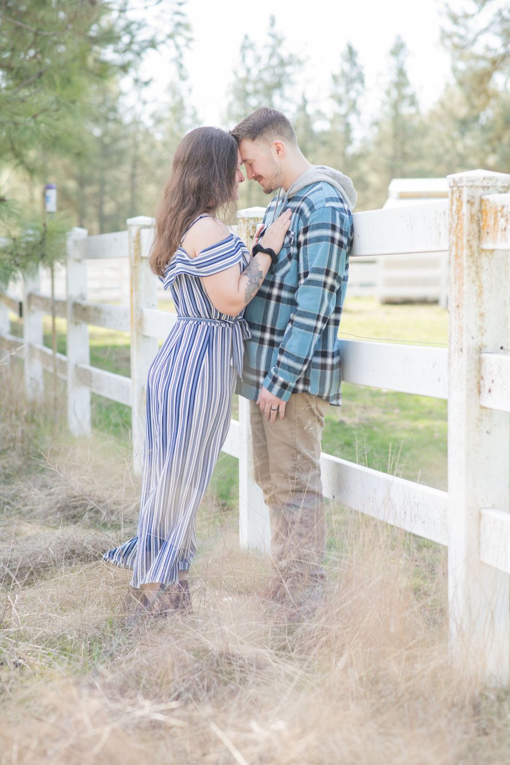A couple shares an intimate nose-to-nose moment leaning against a white fence in a pine forest.