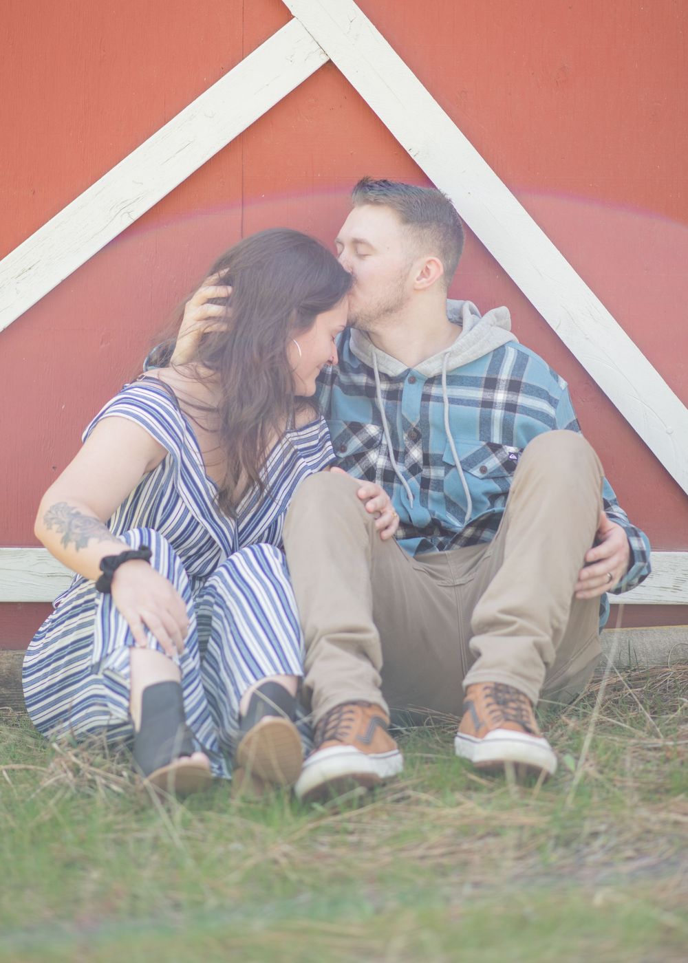 A couple sits together kissing against a red barn wall in a playful outdoor photo session.