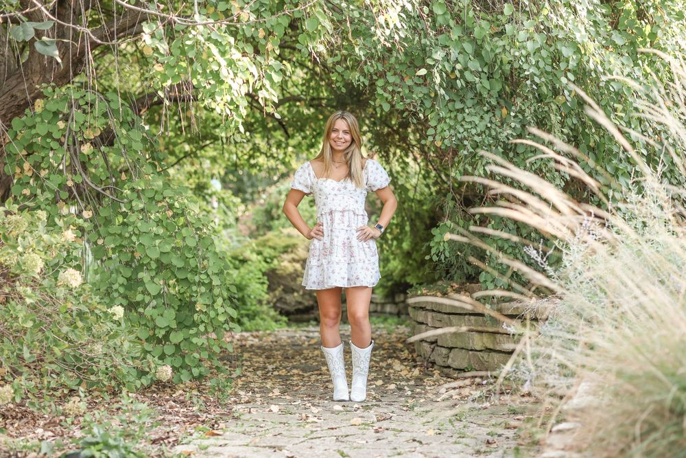 A full-length portrait on a garden path wearing a white floral dress and boots.