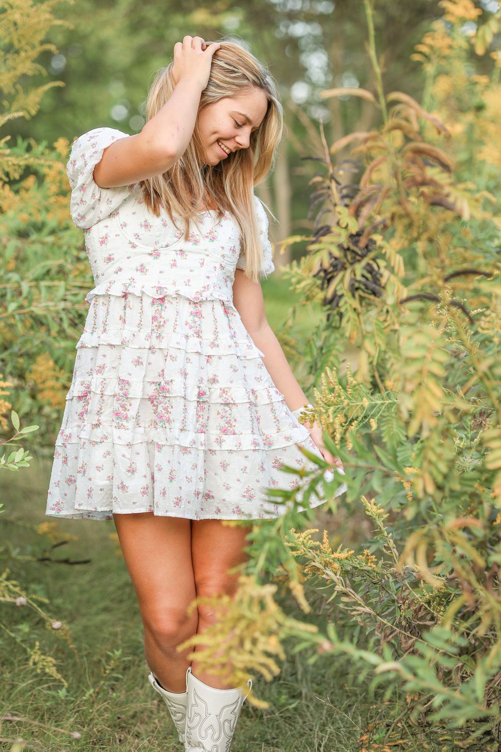 Playful pose in a short white floral dress and white boots surrounded by autumn plants.