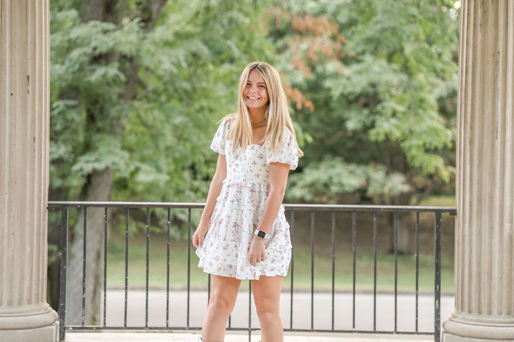 A floral white sundress and boots outfit photographed on a columned porch with greenery in the background.