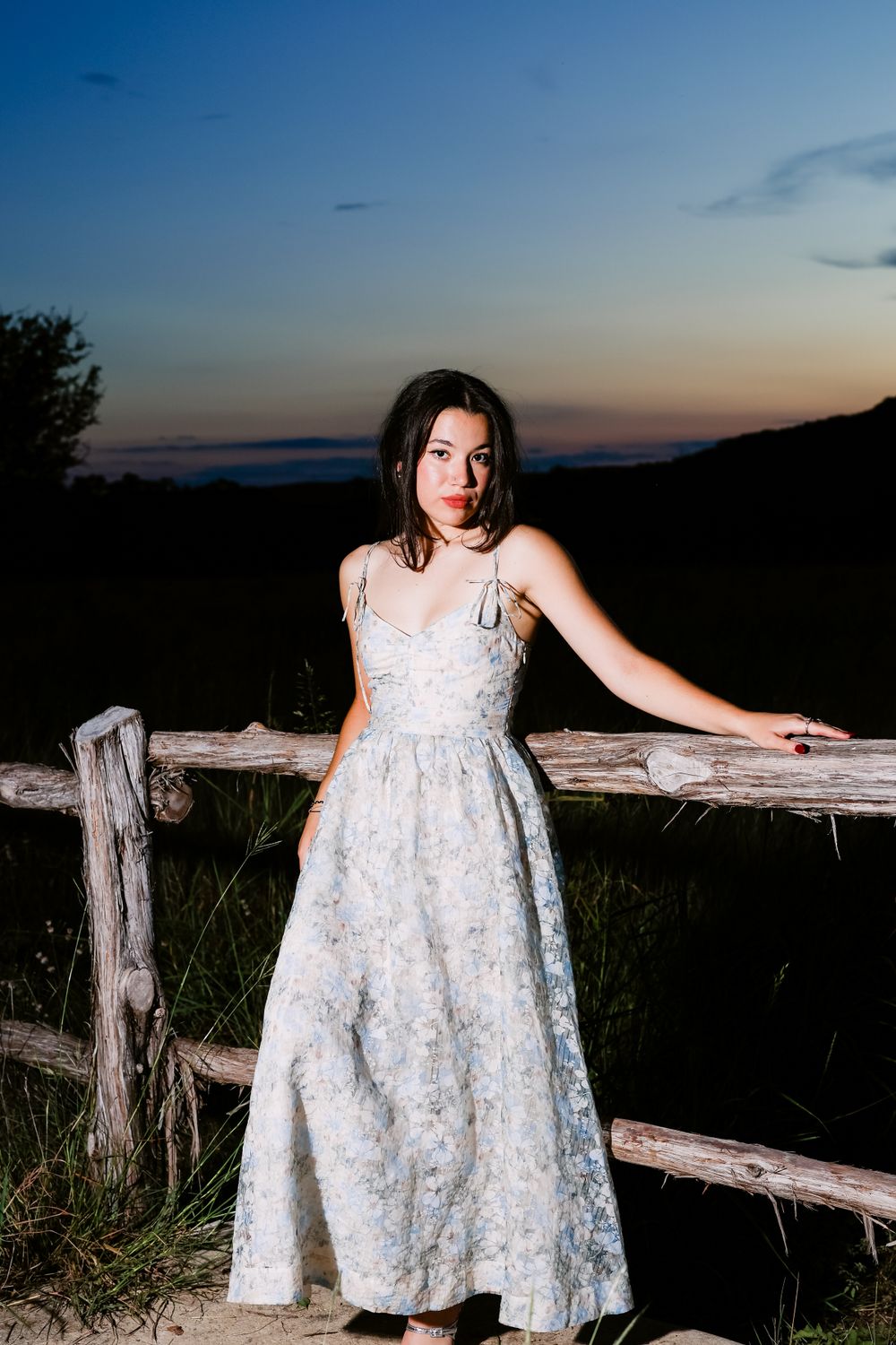 A dramatic sunset portrait by a rustic wooden fence in a flowing white dress.