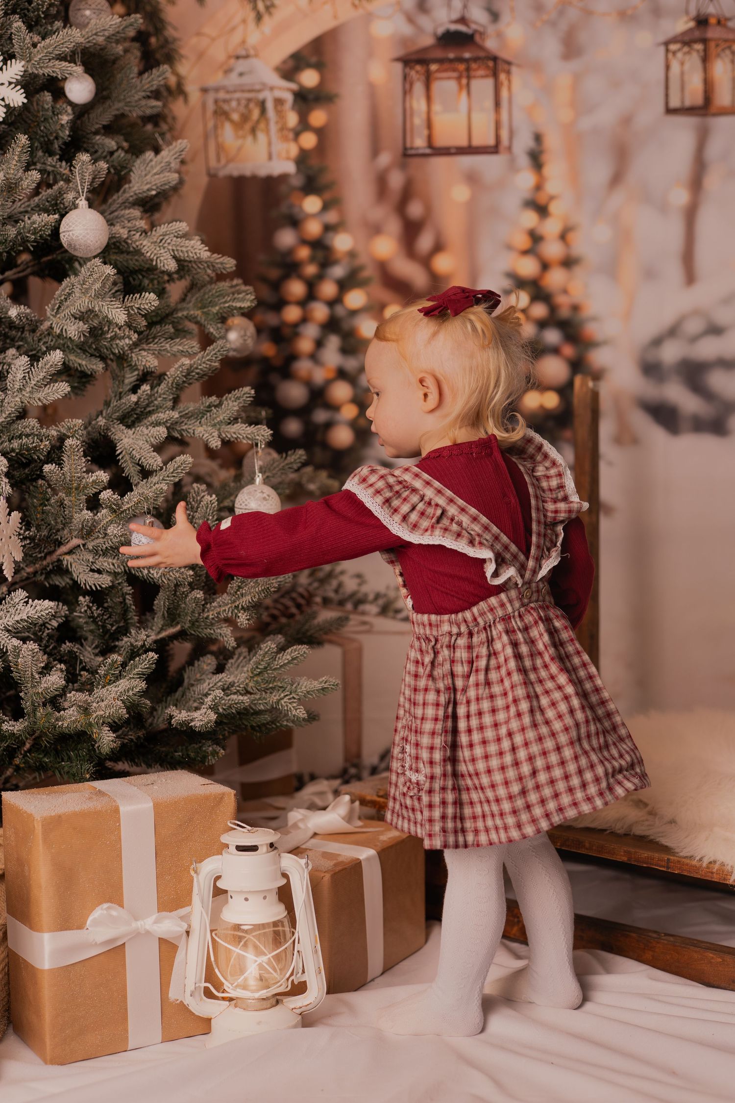 Holiday portrait of a small child in red dress sitting in wicker chair surrounded by Christmas decorations.
