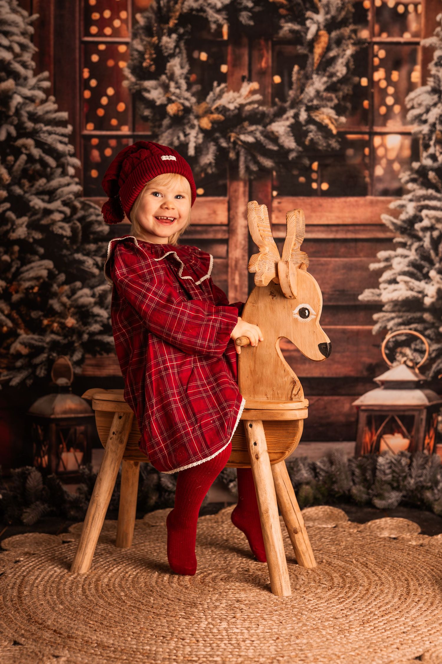 Holiday portrait of a small child in red dress sitting in wicker chair surrounded by Christmas decorations.