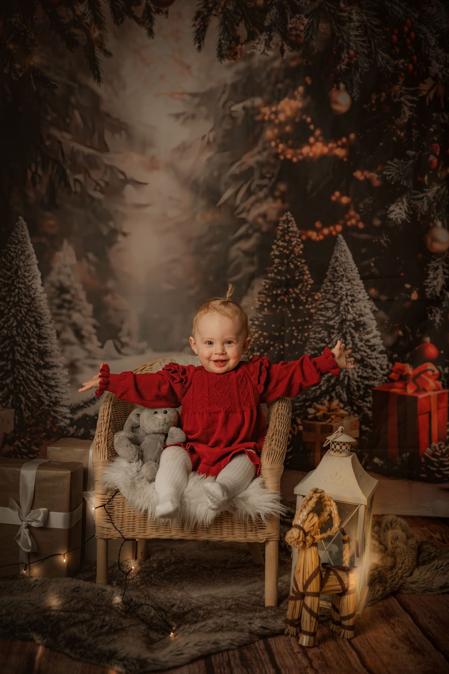 Holiday portrait of a small child in red dress sitting in wicker chair surrounded by Christmas decorations.