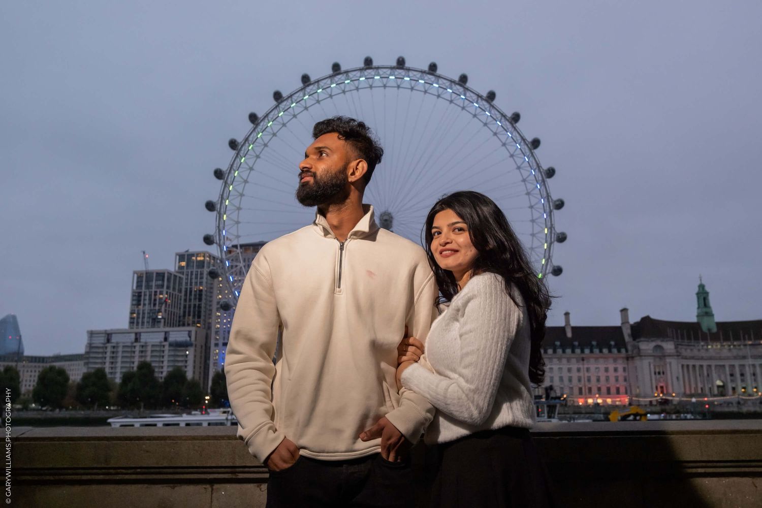 A couple shares an intimate moment in front of the London Eye at twilight.