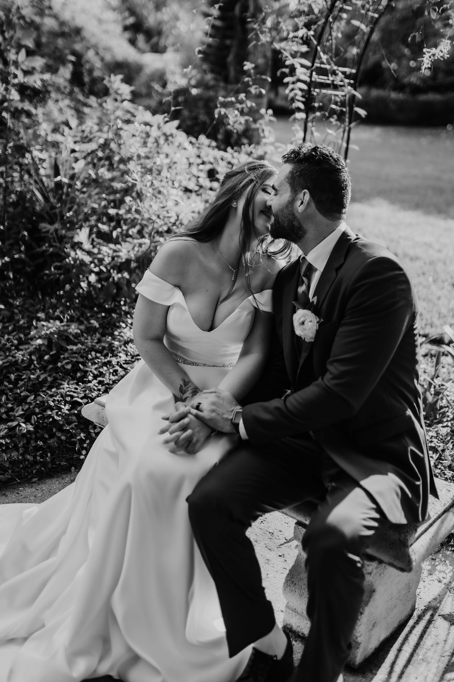 Wedding couple embraces in a sunlit garden with white floral bouquet at their feet.