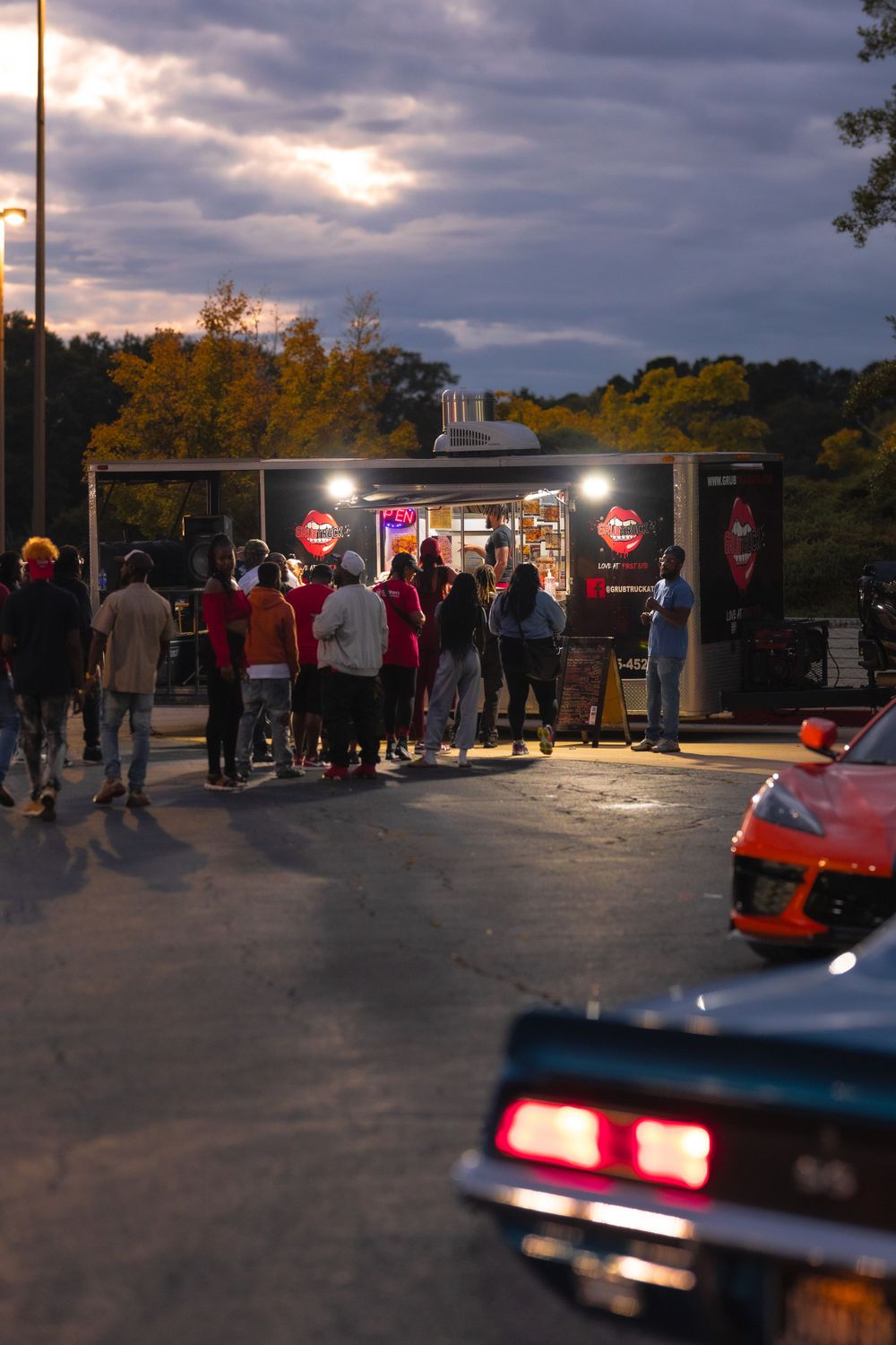 People lined up at food truck during sunset with classic cars visible in background. | Shot By Lo™
