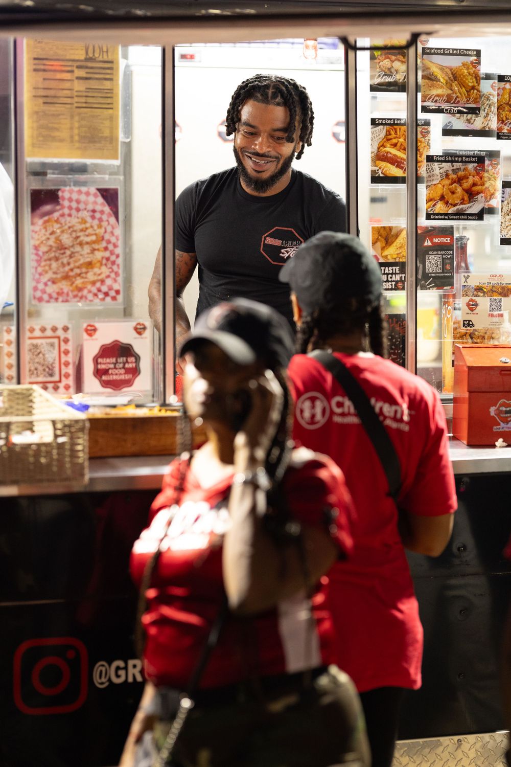 Food vendor in black shirt serving customers at night through illuminated service window. | Shot By Lo™