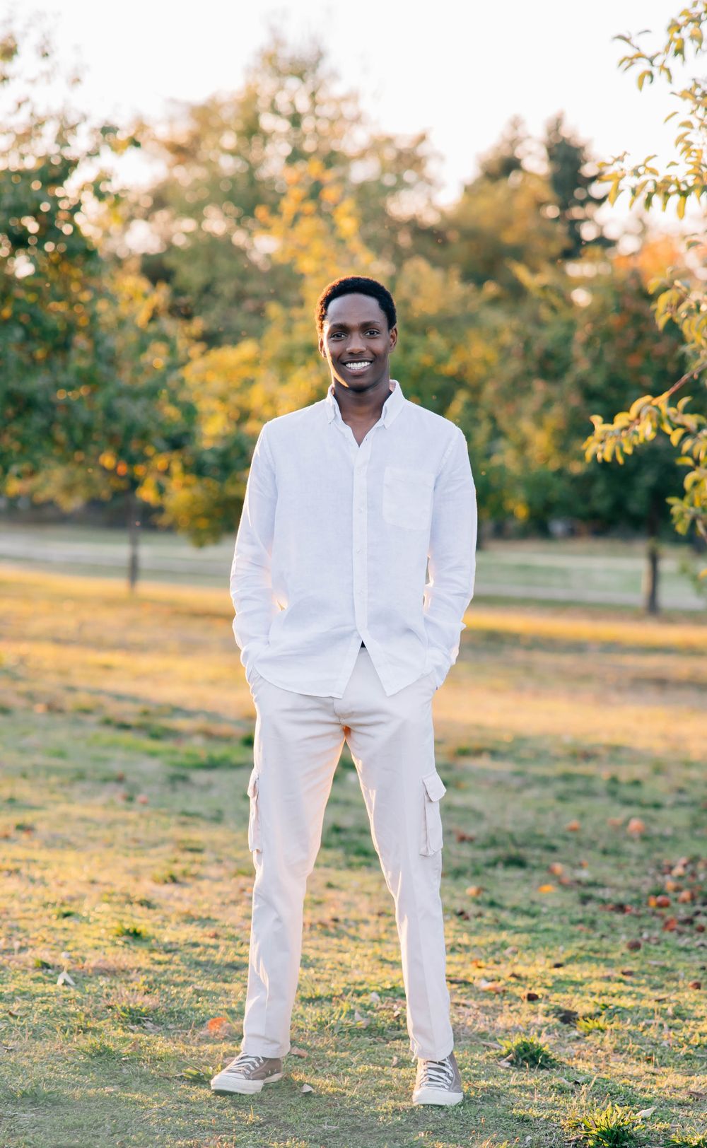 Smiling young Black man in white outfit standing in a sunny park with autumn trees.