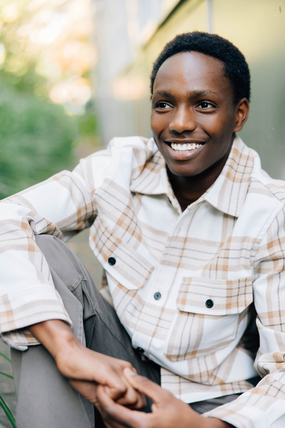Cheerful young Black man in plaid shirt sitting outdoors smiling in natural light setting.