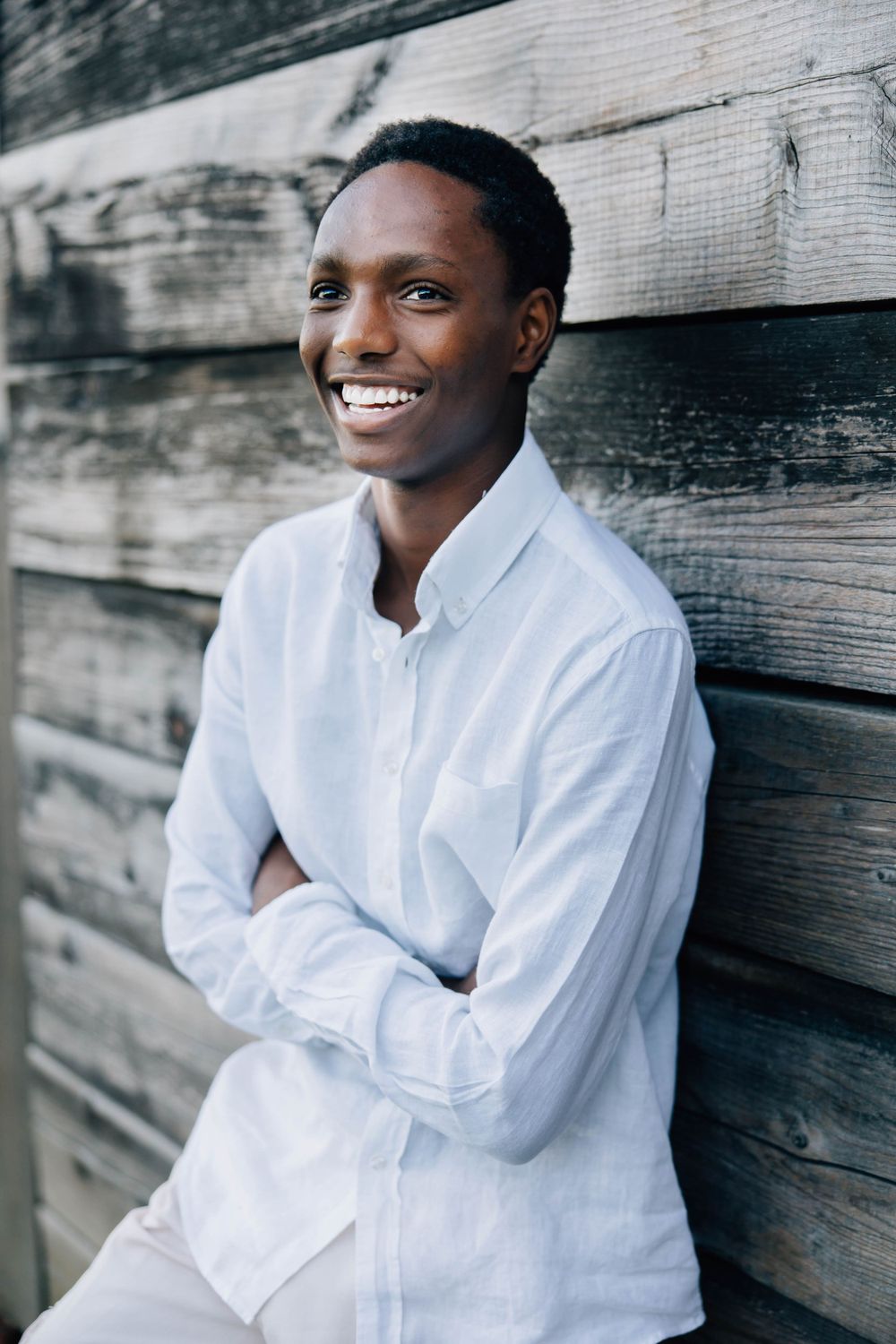 Happy young Black man in light blue shirt leaning against wooden wall with arms crossed.