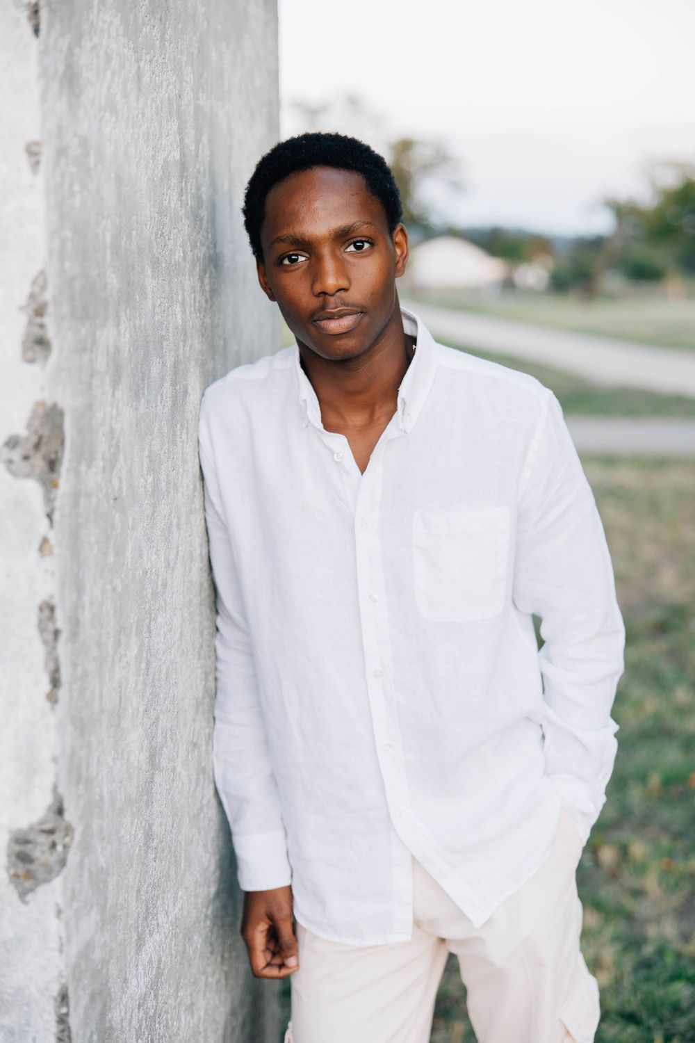 Young Black man in white shirt leaning against concrete wall with a calm expression outdoors.