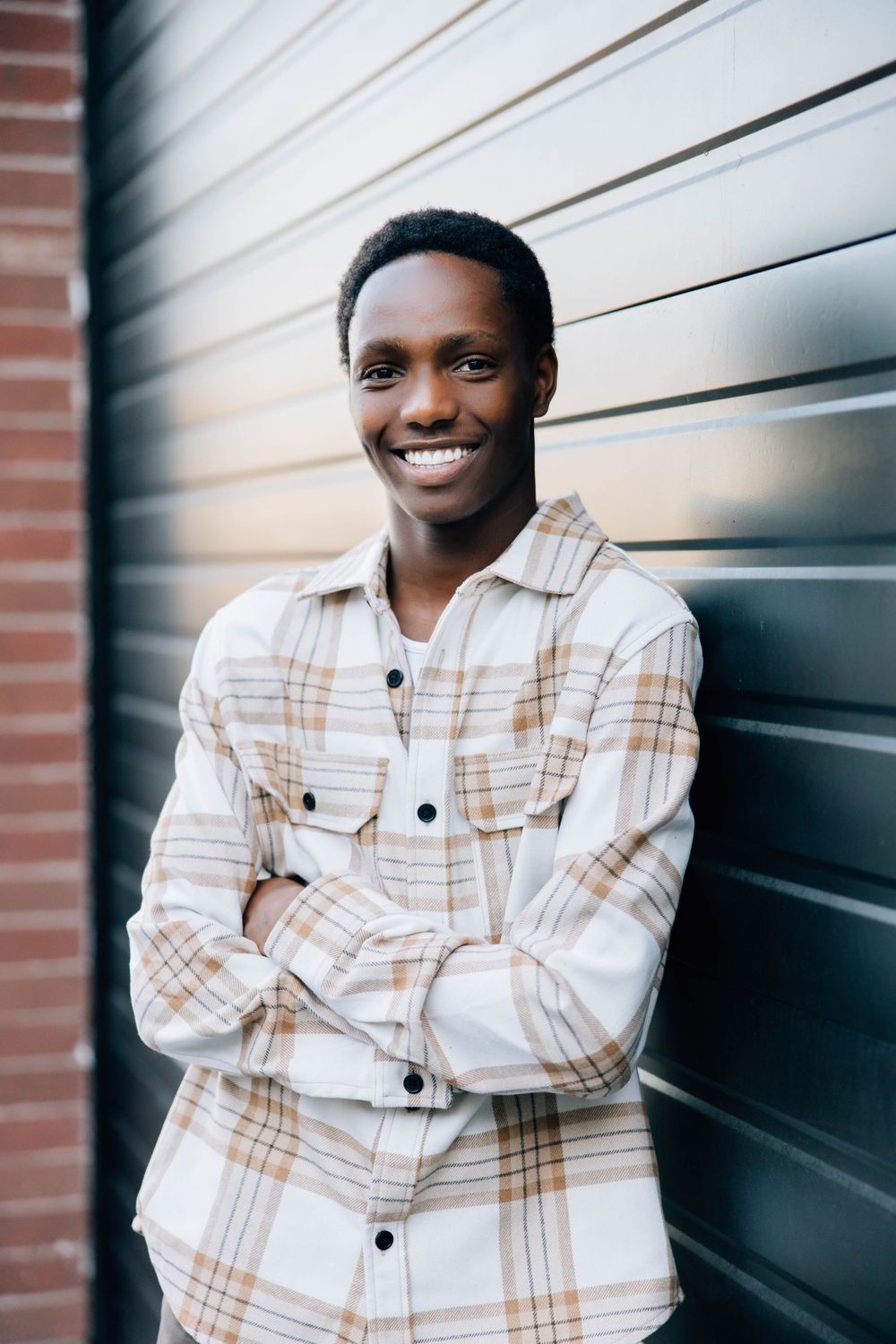 Confident young Black man in plaid flannel shirt smiling with arms crossed against dark wall.