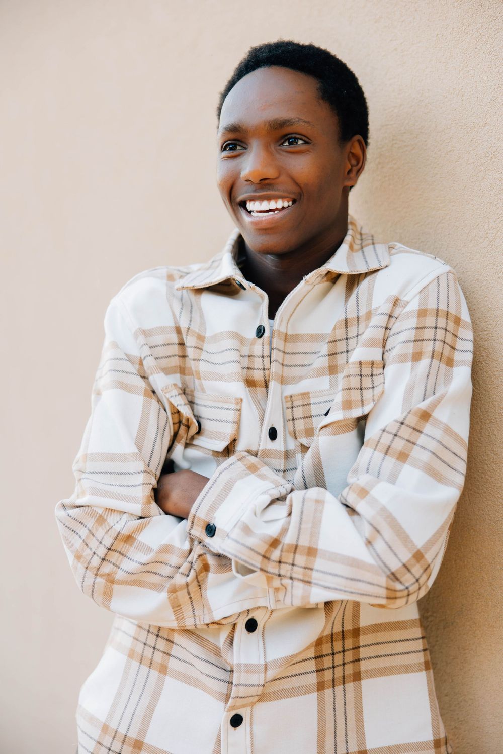 Smiling young Black man in plaid flannel shirt with arms crossed leaning against beige wall.