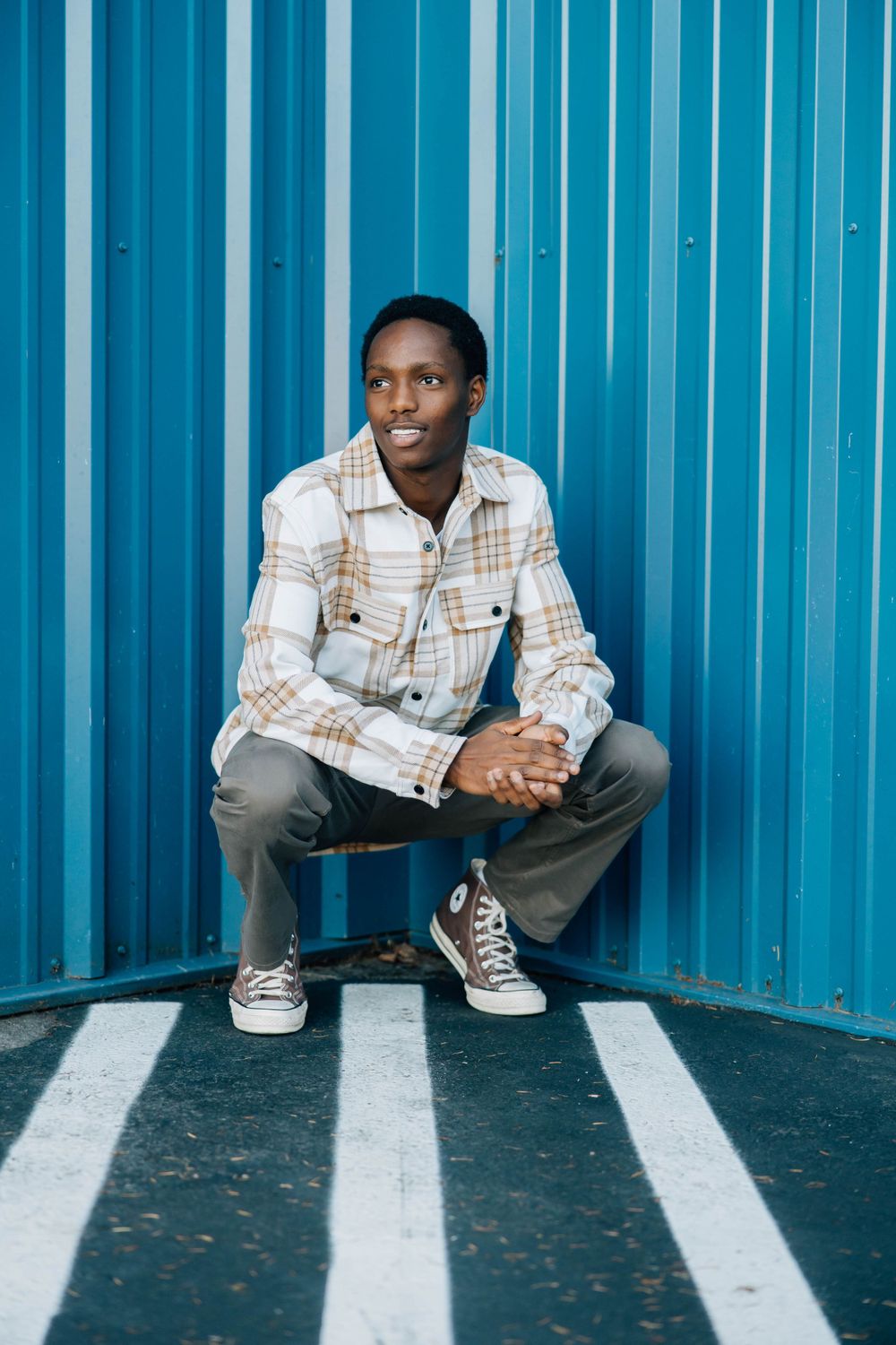 Young Black man in plaid jacket crouching confidently against a bright blue metal wall.