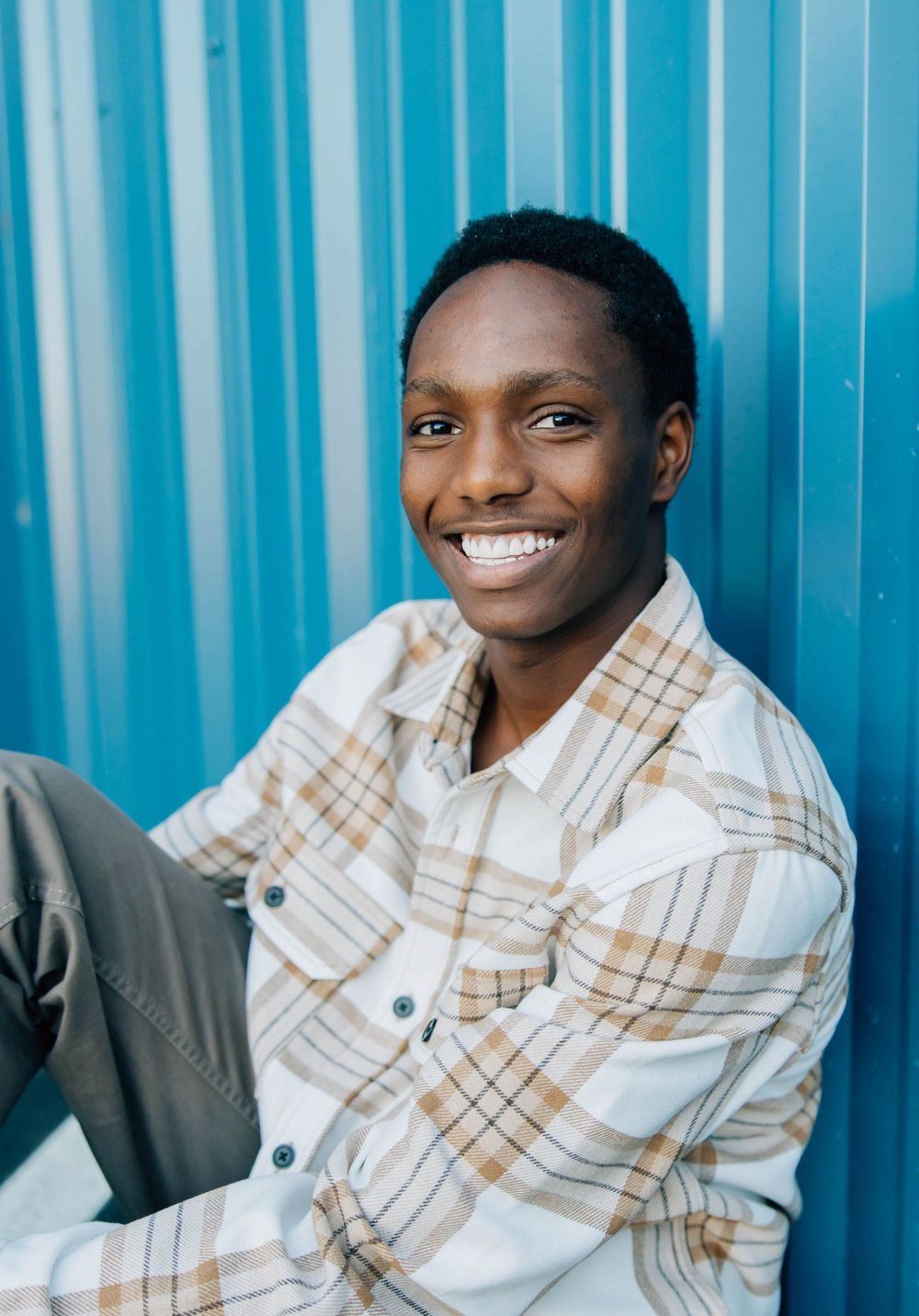 Smiling young Black man in plaid shirt sitting casually against a vibrant blue metal wall.