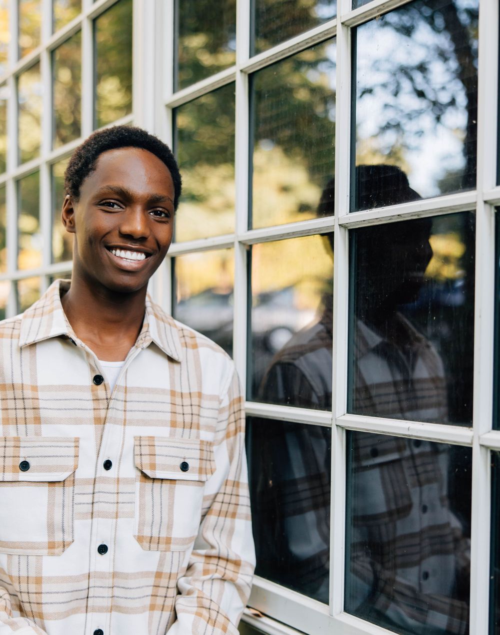 Happy young Black man in plaid shirt smiling beside large window with reflection visible.