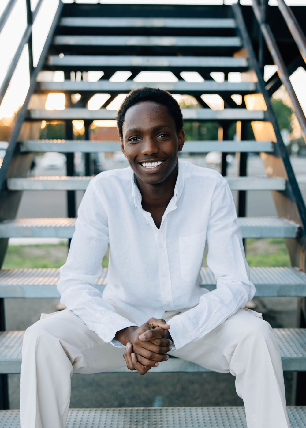 Cheerful young Black man in white outfit sitting on metal stairs smiling outdoors at sunset.