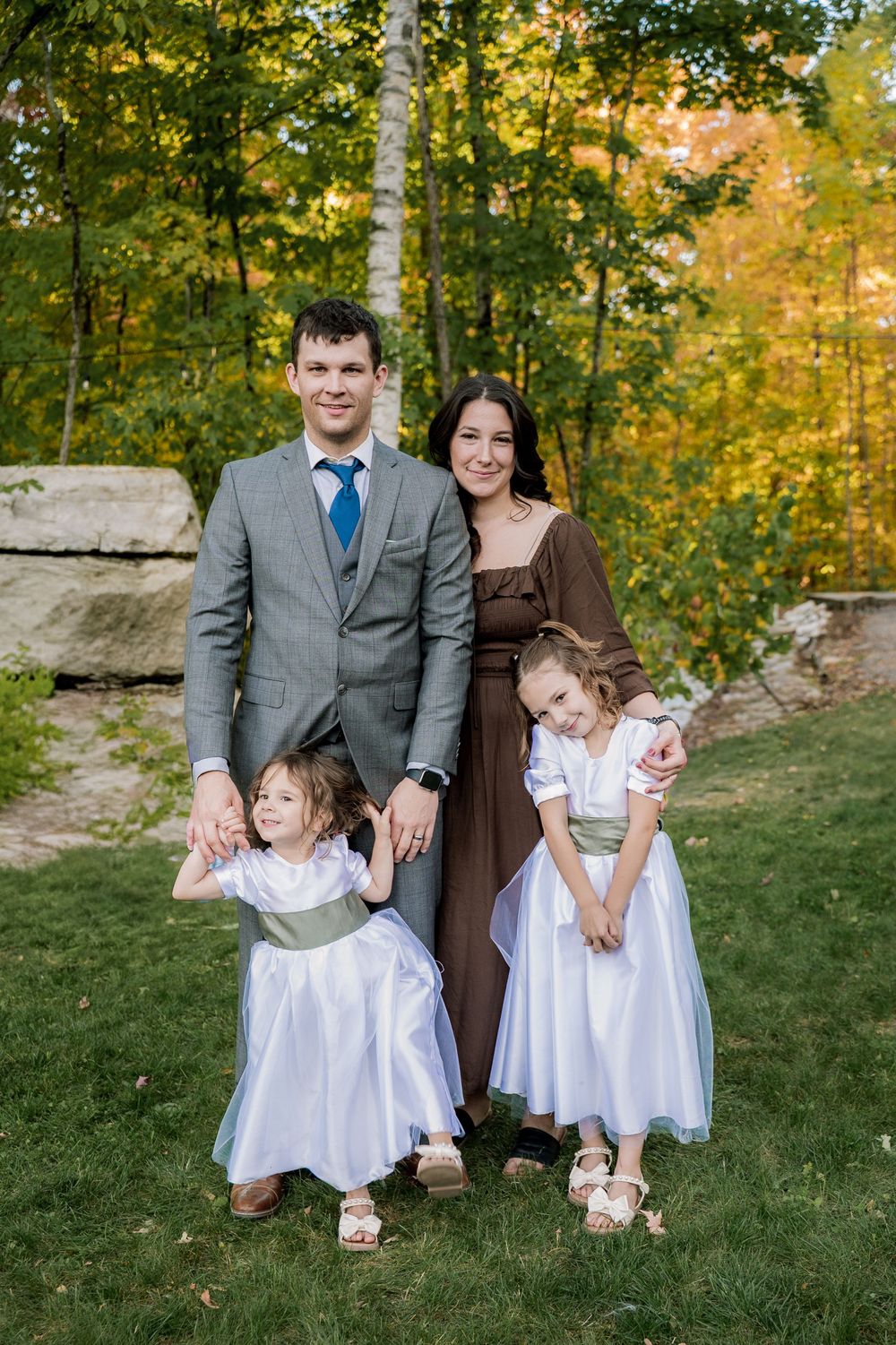 Family sitting on bench in autumn forest setting with young children in white dresses.