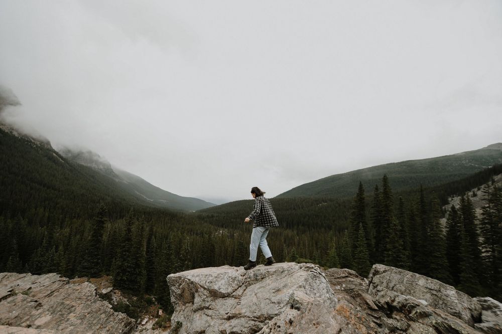 Figure dressed in white stands triumphantly on rocky mountain overlook with misty forest valley below.