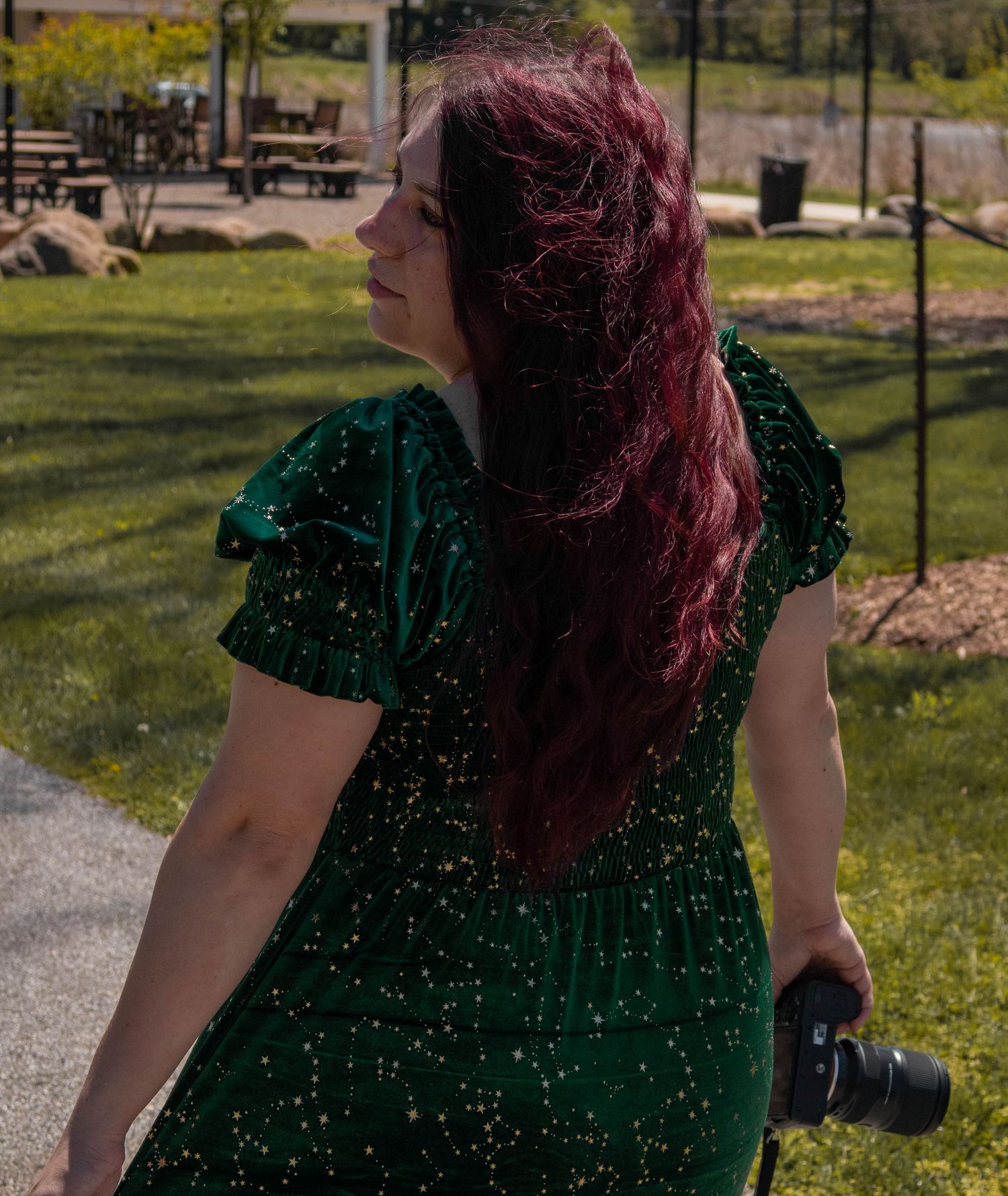 Woman with red hair in green sequin dress holding camera walks through sunny park.