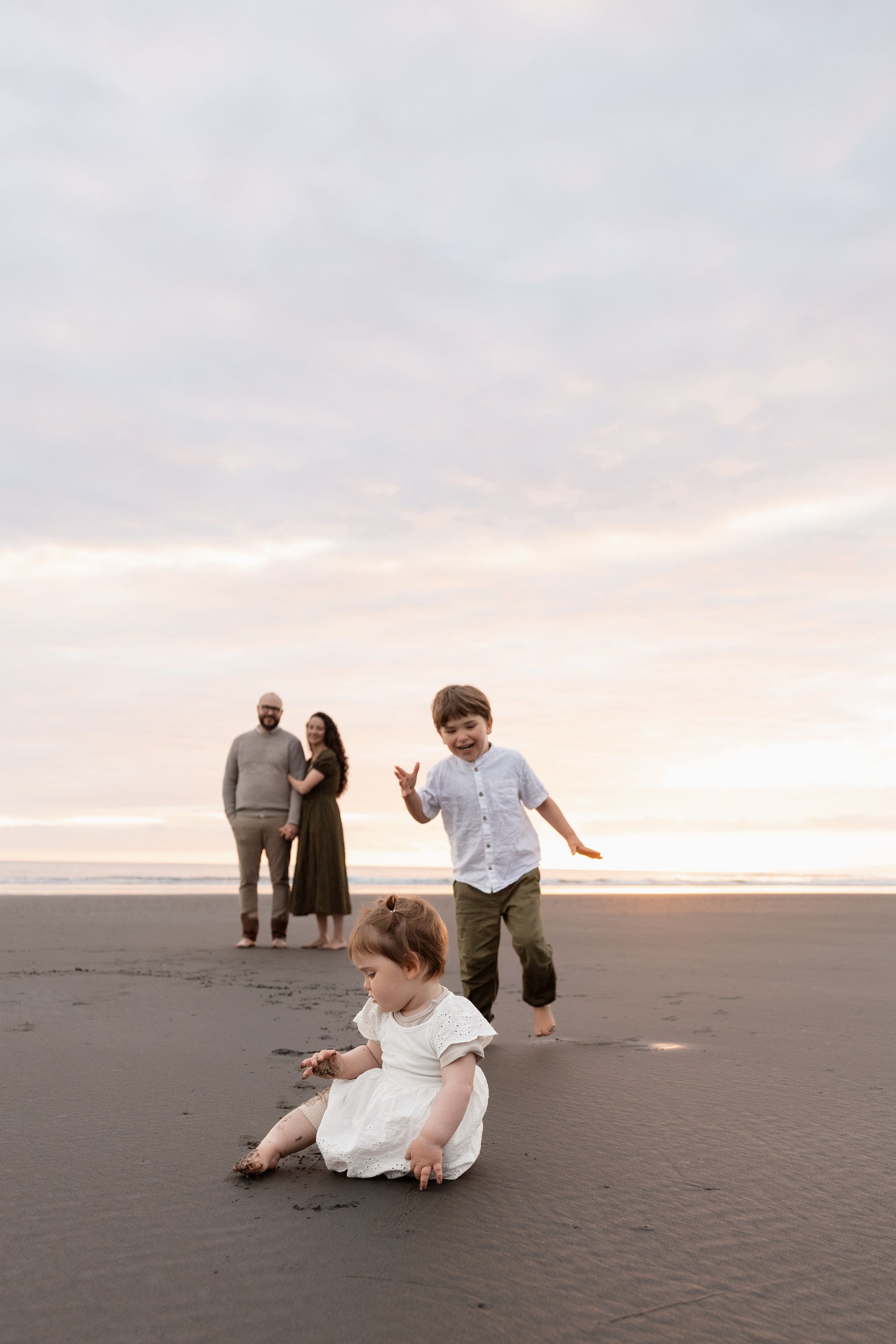 Daughter playing in the sand while brother is running towards her and parents looking, during family beach session Christchurch