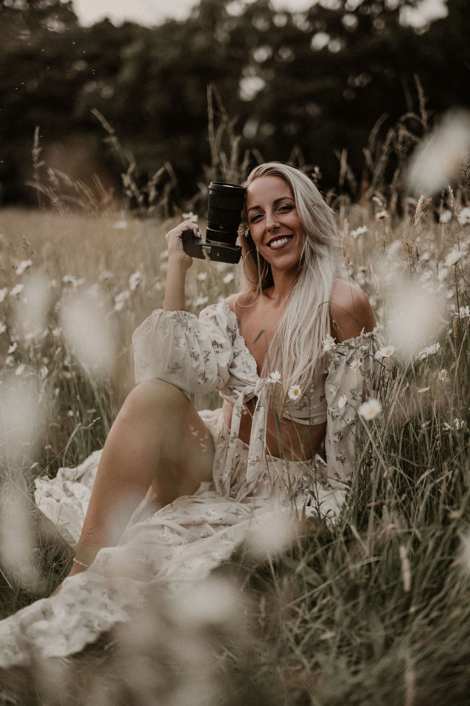 Smiling blonde woman in floral dress sitting in wildflower meadow holding vintage camera outdoors.